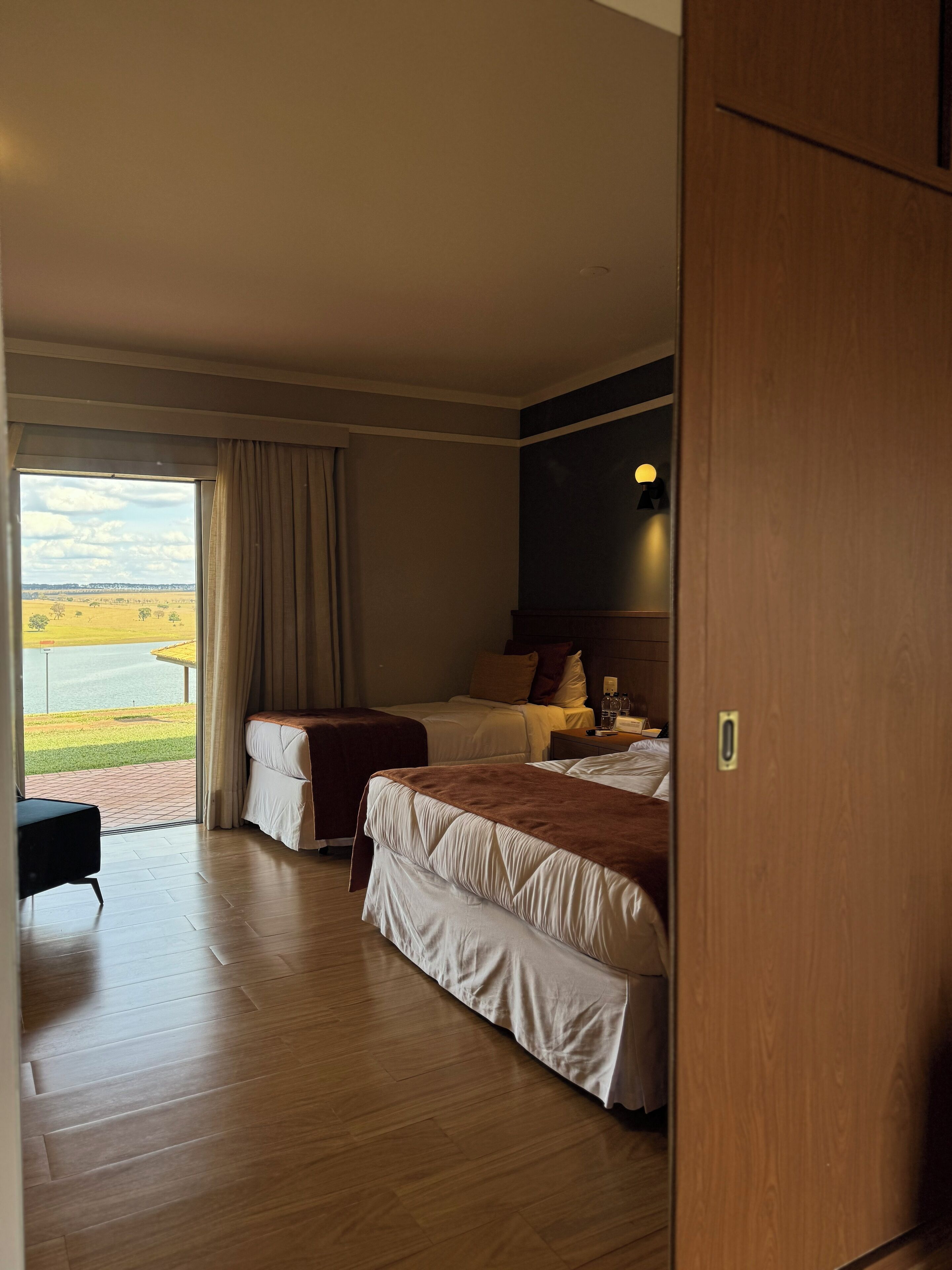 A hotel room with two beds, a wooden floor, a large window with a view of a field and water, and a door to the right.
