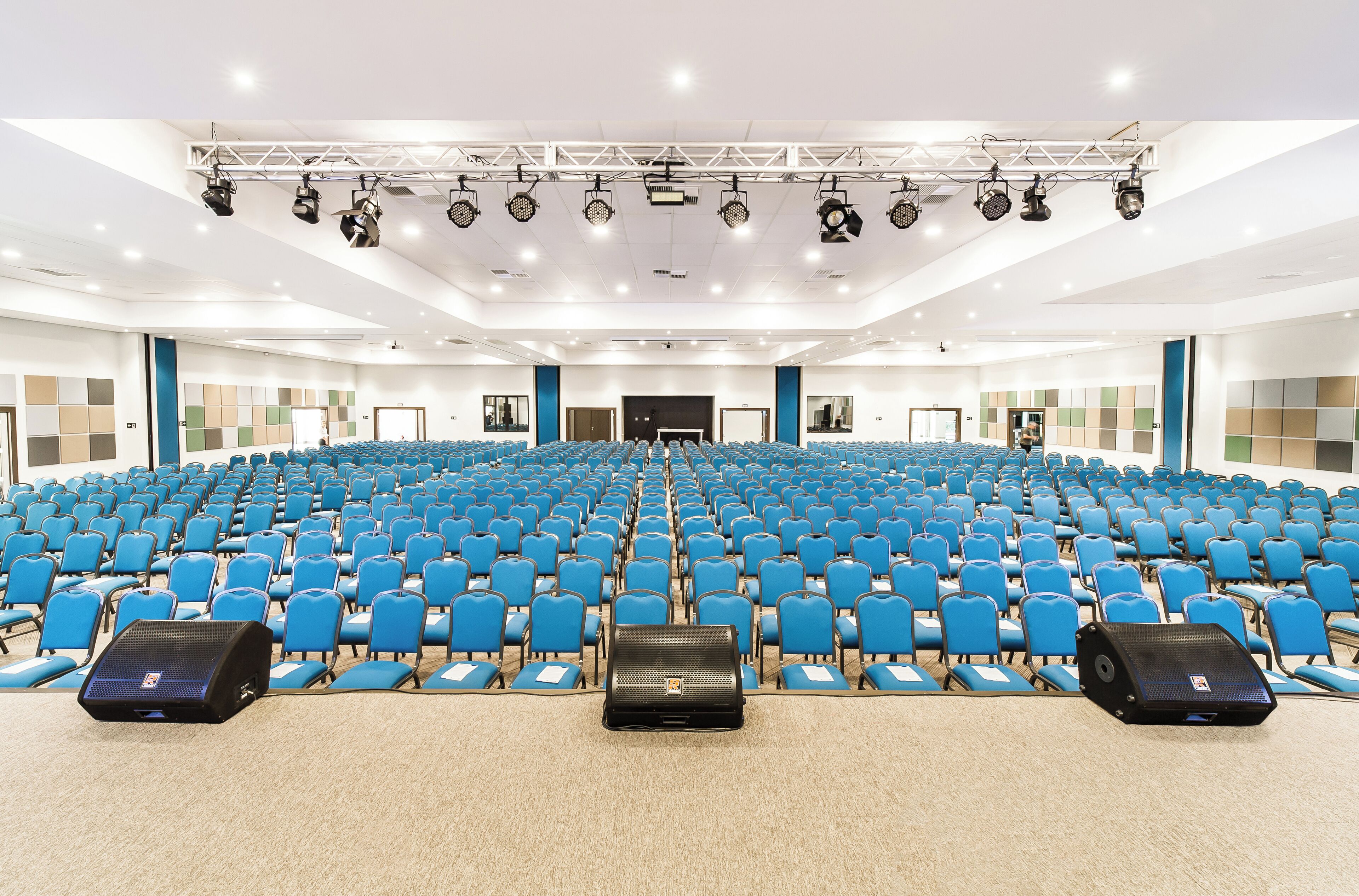 A large auditorium with rows of blue chairs, a stage with lighting equipment, and a black briefcase on the floor.