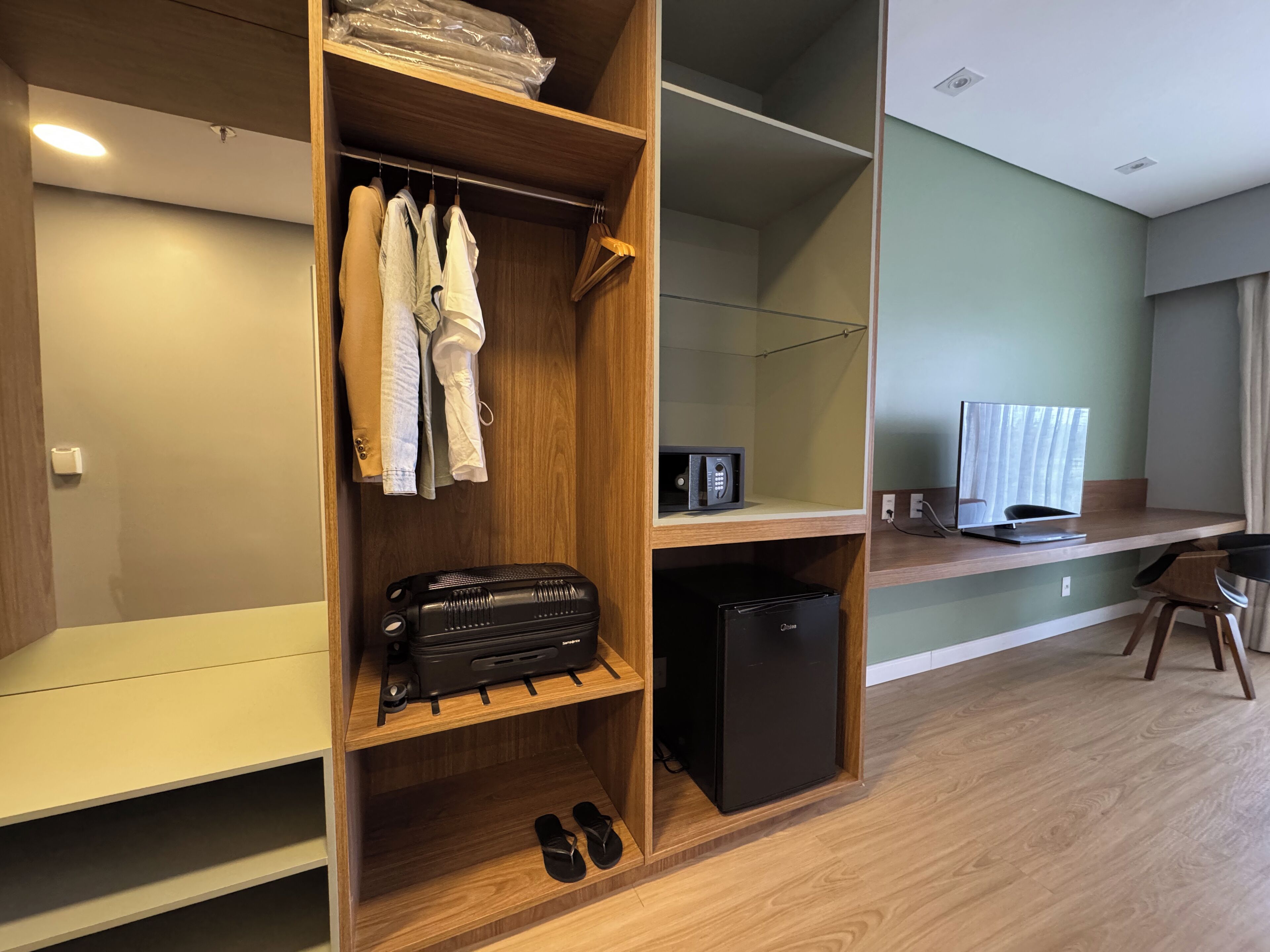 A hotel room with a wooden wardrobe, a desk with a computer, a TV, and a small refrigerator.