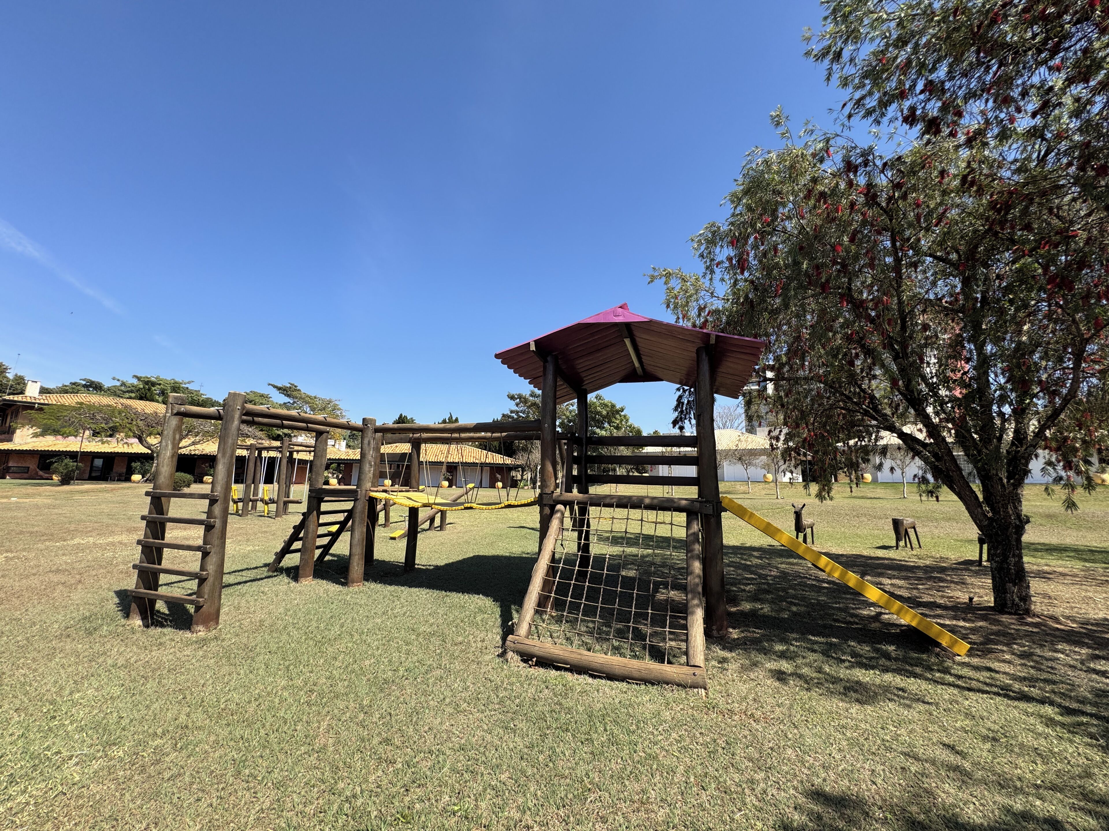 Un parque infantil con una estructura de madera para escalar, un tobogán amarillo y una zona delimitada por una valla.