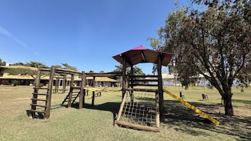 Un parque infantil con una estructura de madera para escalar, un tobogán amarillo y una zona delimitada por una valla.