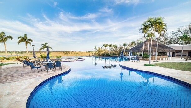A swimming pool with lounge chairs and umbrellas, surrounded by palm trees and a building with a thatched roof. - Ecoblue Resort Avaré (Avaré)