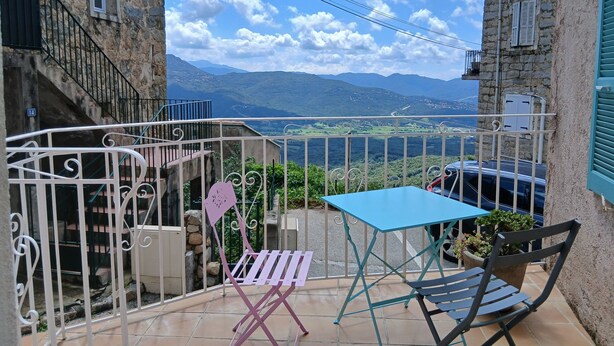 Un balcón con vistas a montañas, una mesa azul, una silla rosa y una silla negra.