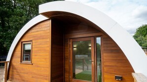 A modern, curved wooden structure with a glass door and window, surrounded by a deck and green grass.
