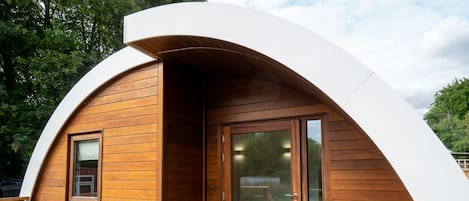 A modern, curved wooden structure with a glass door and window, surrounded by a deck and green grass.
