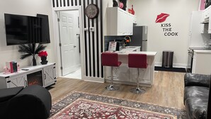 Un salon moderne avec un téléviseur, une cheminée, un tapis rouge et un coin cuisine agrémenté d’une horloge et d’une pancarte indiquant « EMBRASSEZ LE CÉLÉBRATEUR ».