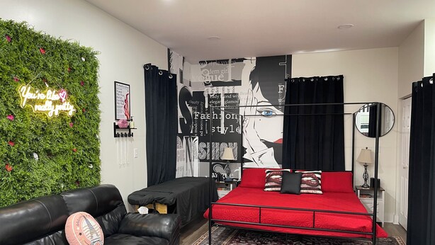 Une chambre moderne avec un lit rouge, des rideaux noirs, un mur vert orné d’un néon, et un canapé en cuir noir.