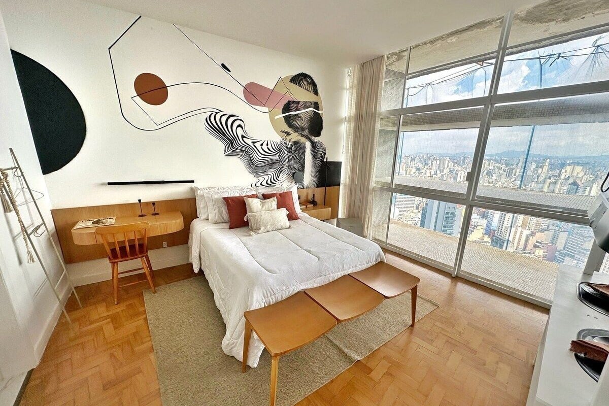 Une chambre moderne avec un grand lit, un bureau et une vue sur le paysage urbain, grâce à de grandes fenêtres.