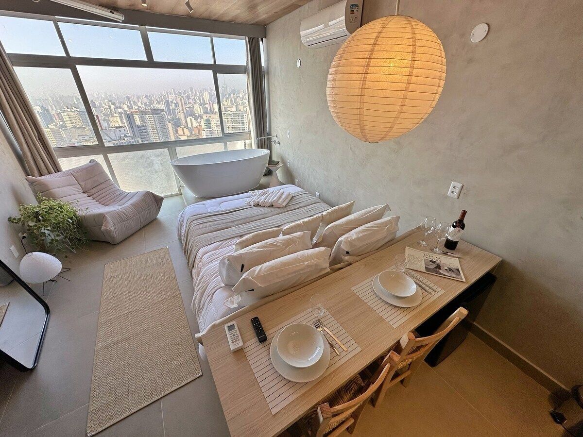 Une chambre d’hôtel moderne avec vue sur la ville, un grand lit, une table à manger dressée avec des assiettes, des verres et une bouteille de vin, et une grande lanterne en papier suspendue au plafond.