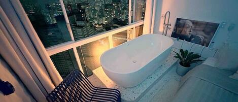 A modern bathroom with a freestanding bathtub, a large window with a city view, and a striped chair.