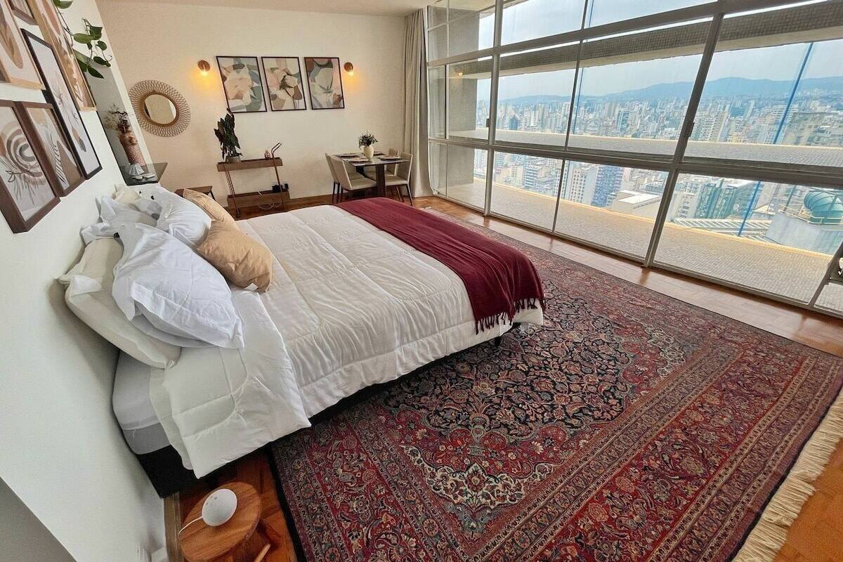 Une chambre avec un grand lit, un tapis rouge à motifs, un bureau avec une chaise et une vue sur la ville à travers de grandes fenêtres.