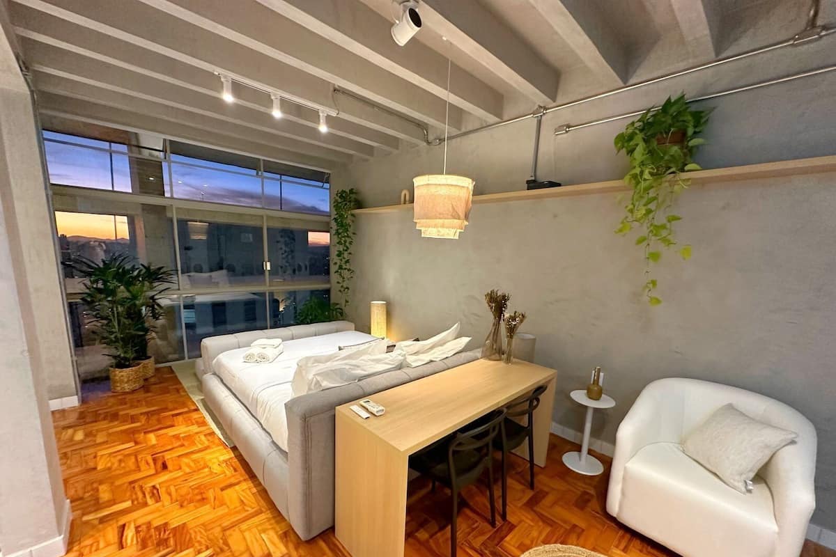 Une chambre moderne avec un lit, un bureau, une chaise et un canapé. On y trouve également des plantes et une fenêtre donnant sur un beau paysage.