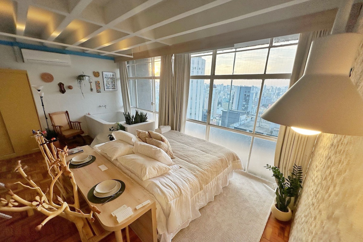 Une chambre moderne dotée d’un grand lit, d’un coin salon avec une table et offrant une vue sur la ville grâce à de grandes fenêtres.