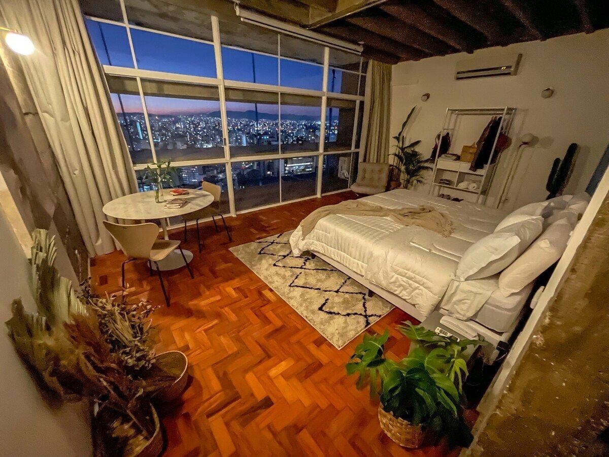 Un dormitorio con un ventanal grande, una cama, una mesita y una silla.