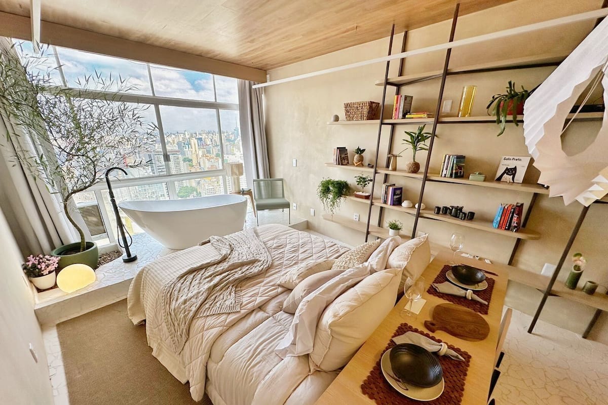Une chambre moderne avec une grande fenêtre, une baignoire, un lit recouvert d’une couette blanche, une table en bois avec des baguettes et une bibliothèque contenant divers objets.