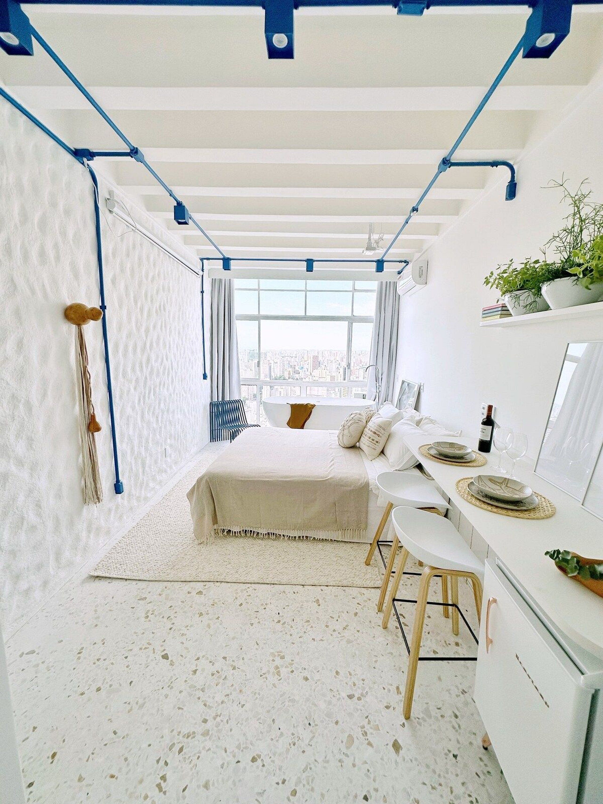 Un dormitorio moderno con una cama, un escritorio y una silla. Se ve una ventana con vistas a la ciudad, y una planta en el escritorio.