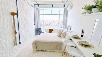 Un dormitorio moderno con una cama, un escritorio y una silla. Se ve una ventana con vistas a la ciudad, y una planta en el escritorio.
