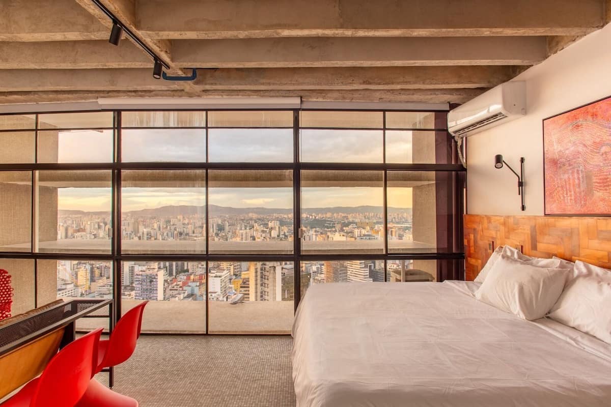 Une chambre d’hôtel dotée d’une grande fenêtre donnant sur la ville, d’un lit, d’un bureau et de chaises rouges.