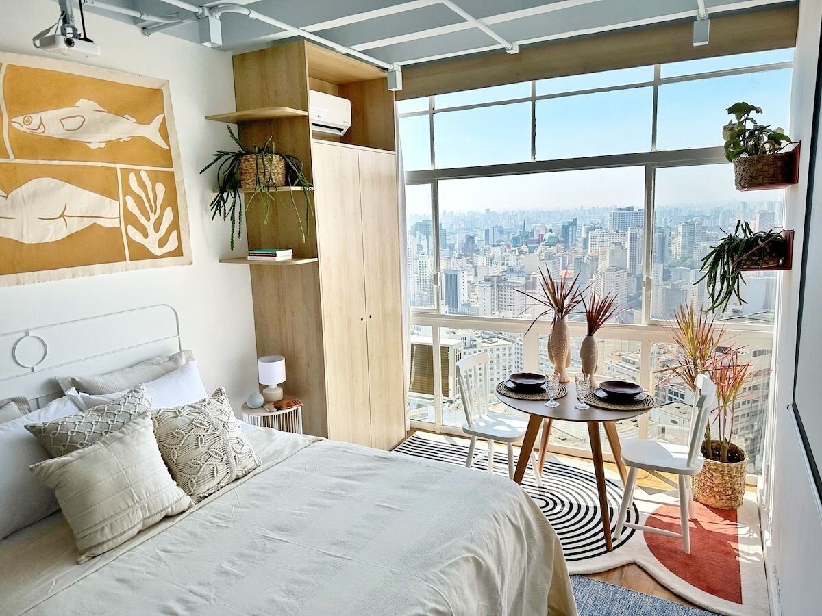 A modern bedroom with a large bed, a city view, a dining area with a table and chairs, and various plants.