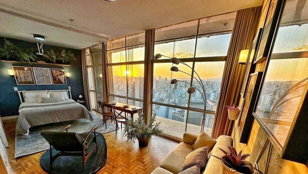 A modern hotel room with a large bed, a sitting area with a sofa and armchair, a dining table, and a city view through large windows. - Vem pro Copan (São Paulo)