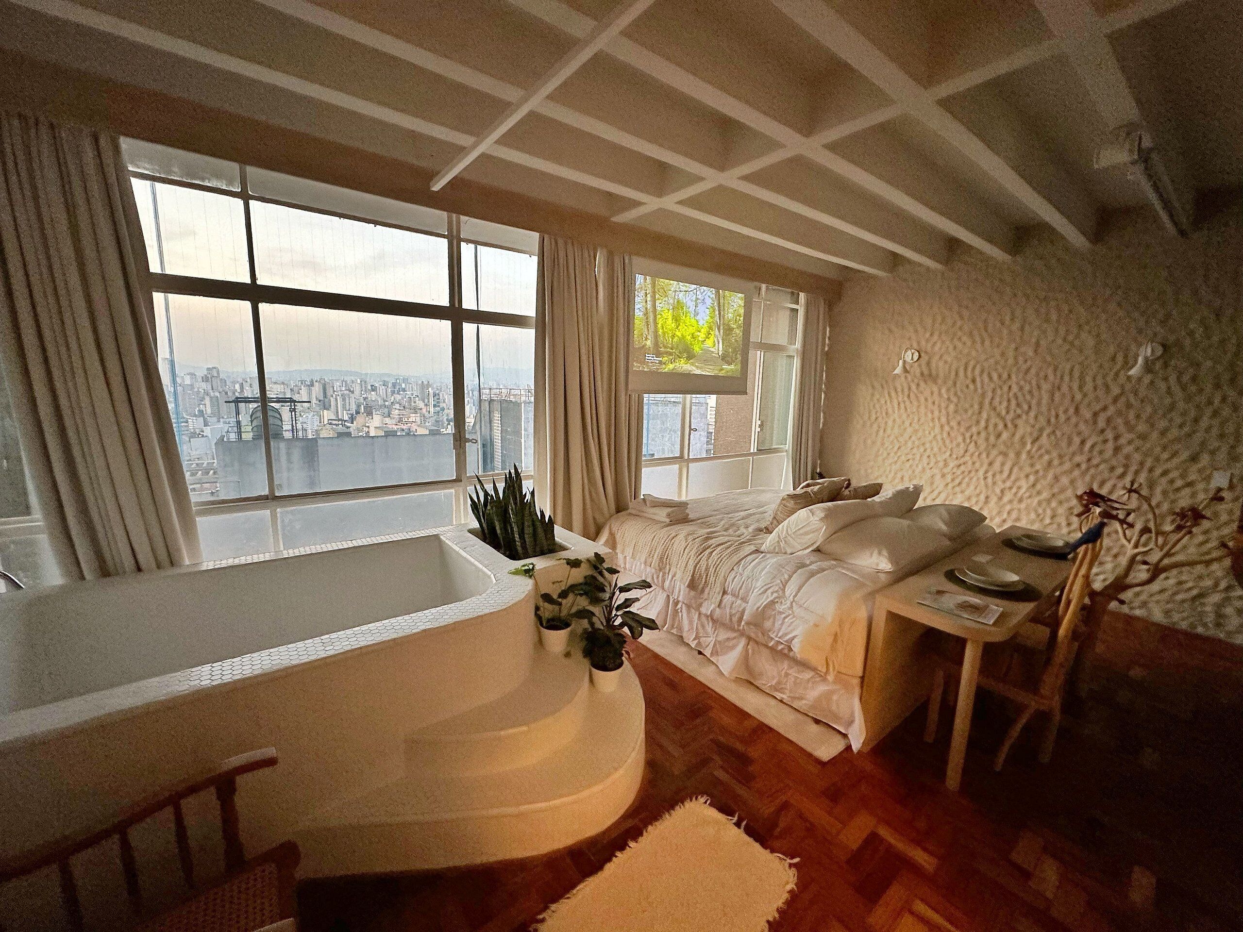 A bedroom with a large window, a bathtub, and a view of a cityscape.