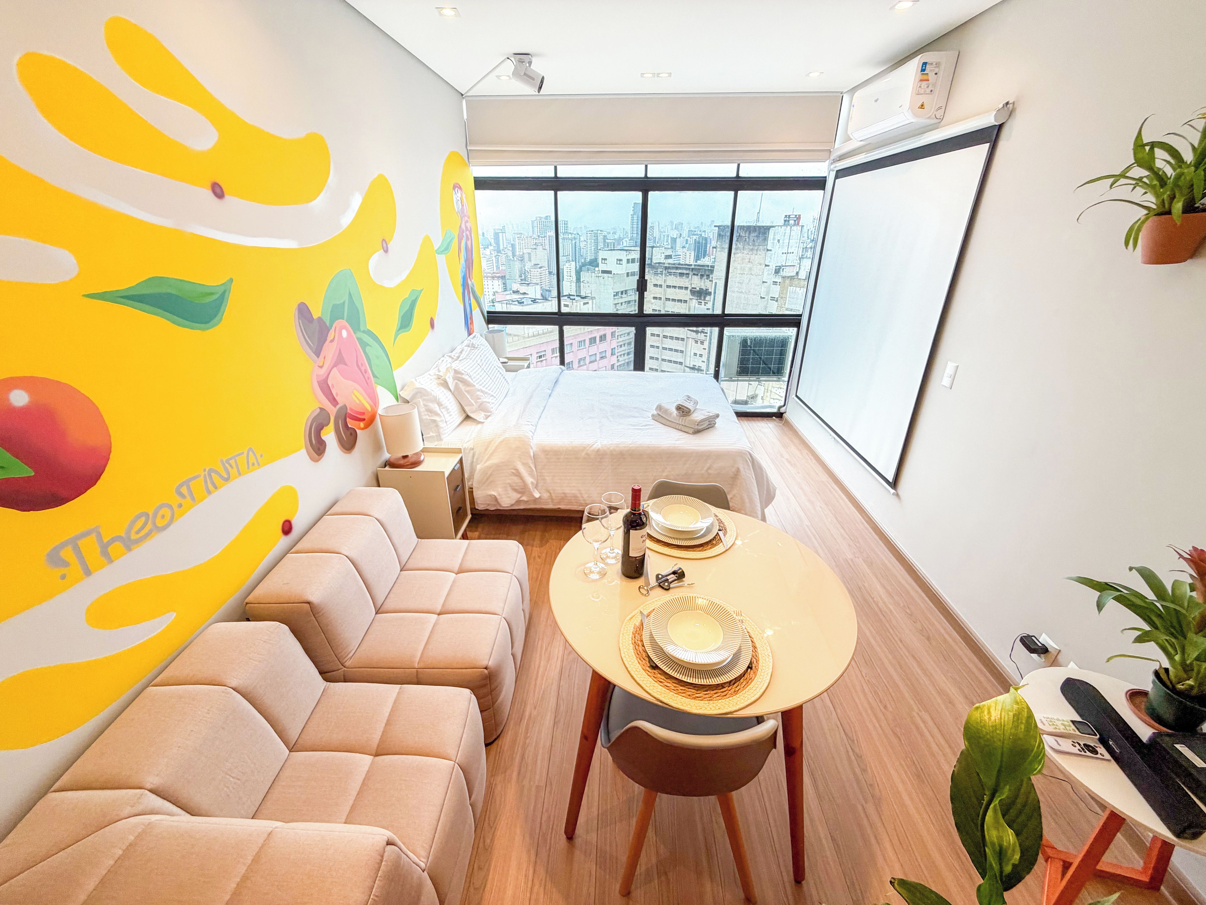 Una moderna camera d'albergo con vista sulla città, un divano, un tavolo da pranzo con piatti e vino, e un murale sulla parete.