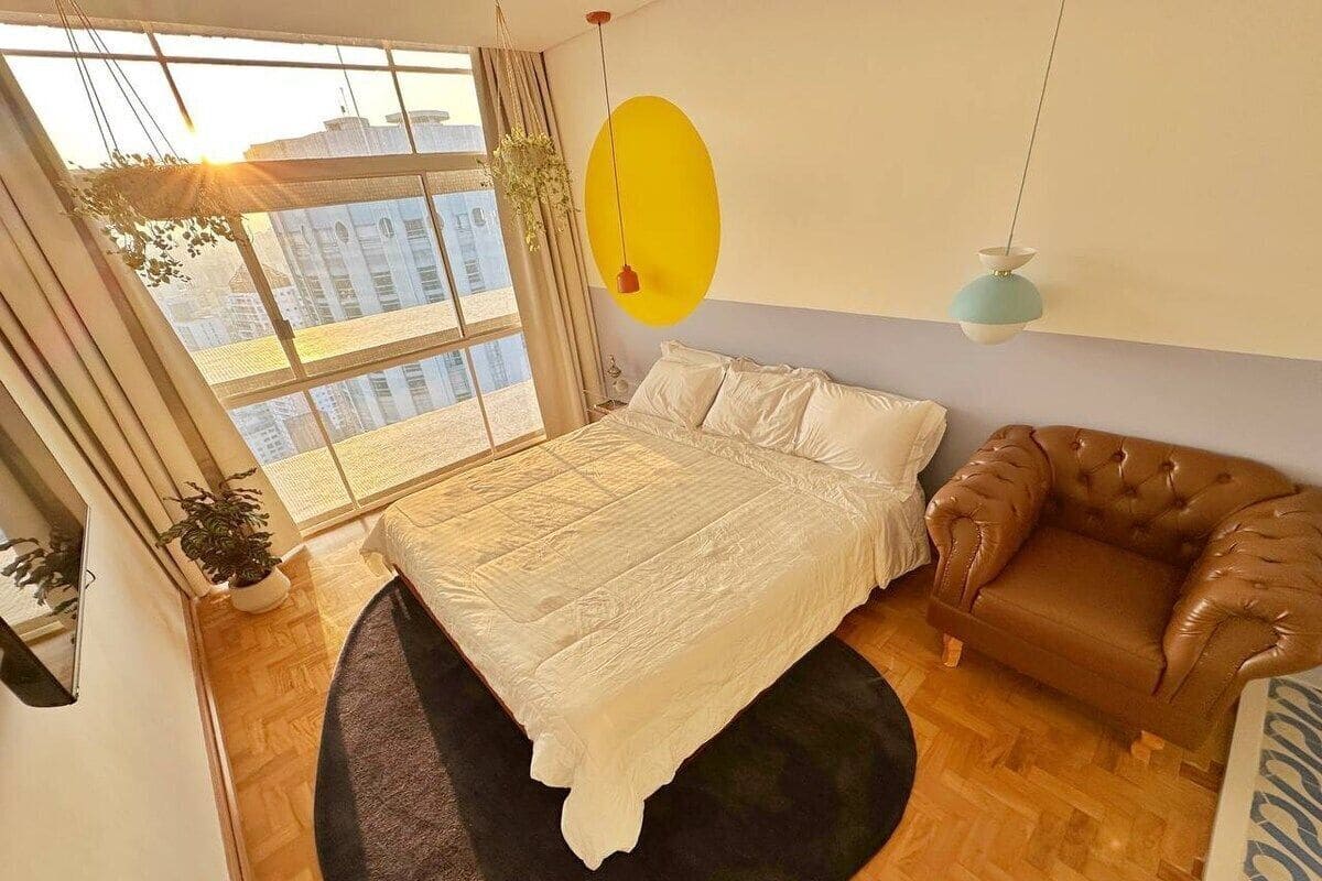 Une chambre avec un lit, un canapé en cuir marron, une grande fenêtre et un élément de décoration jaune sur le mur.