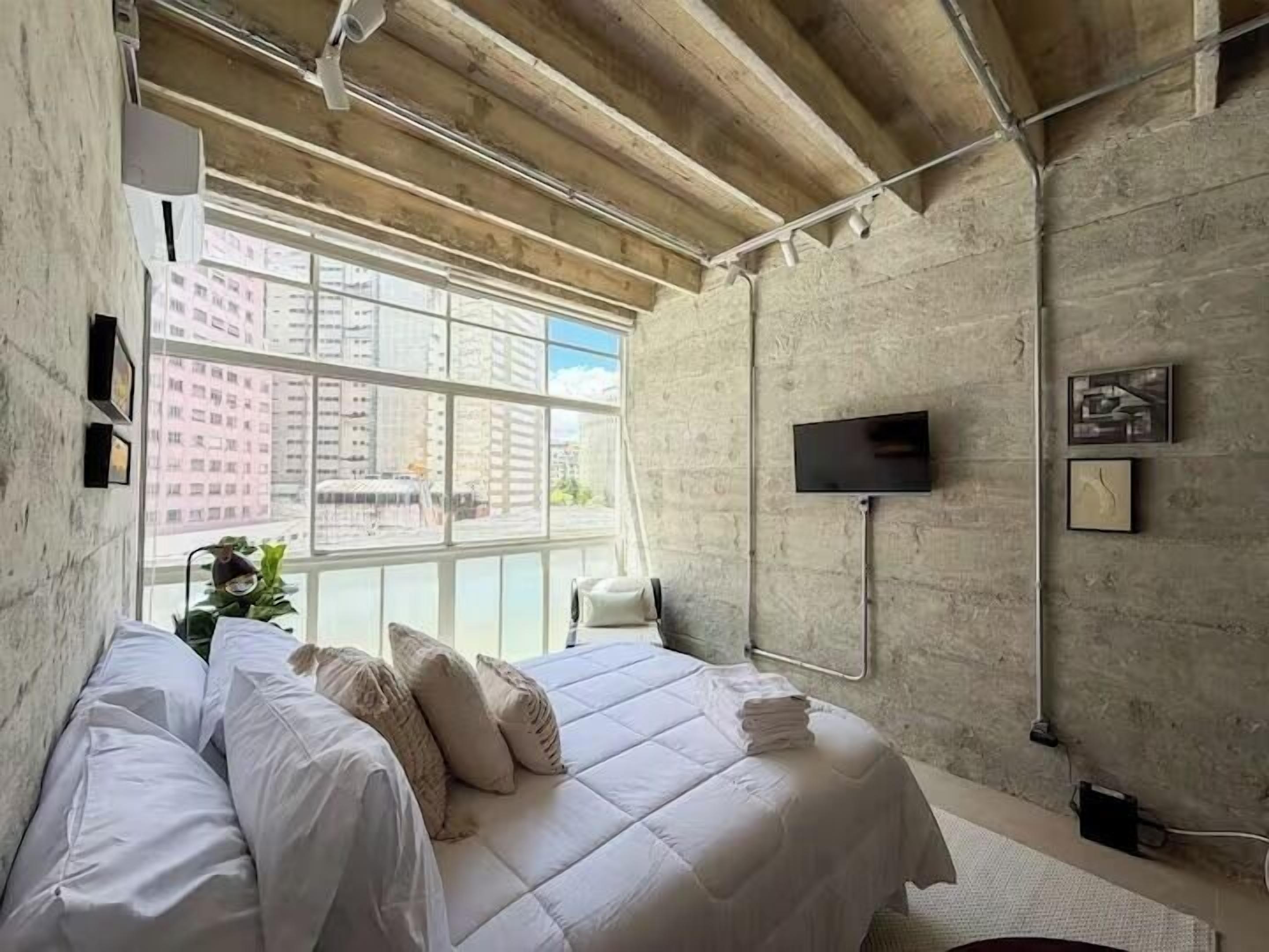 Un dormitorio con una cama grande, un televisor de pantalla plana y vistas a edificios a través de un ventanal amplio.