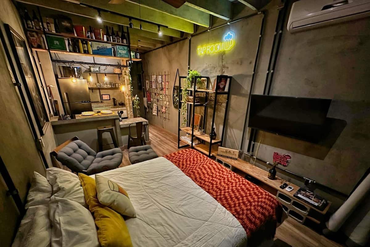 Une chambre d’hôtel compacte comprenant un lit, un petit bureau, un coin salon et un panneau lumineux au néon.