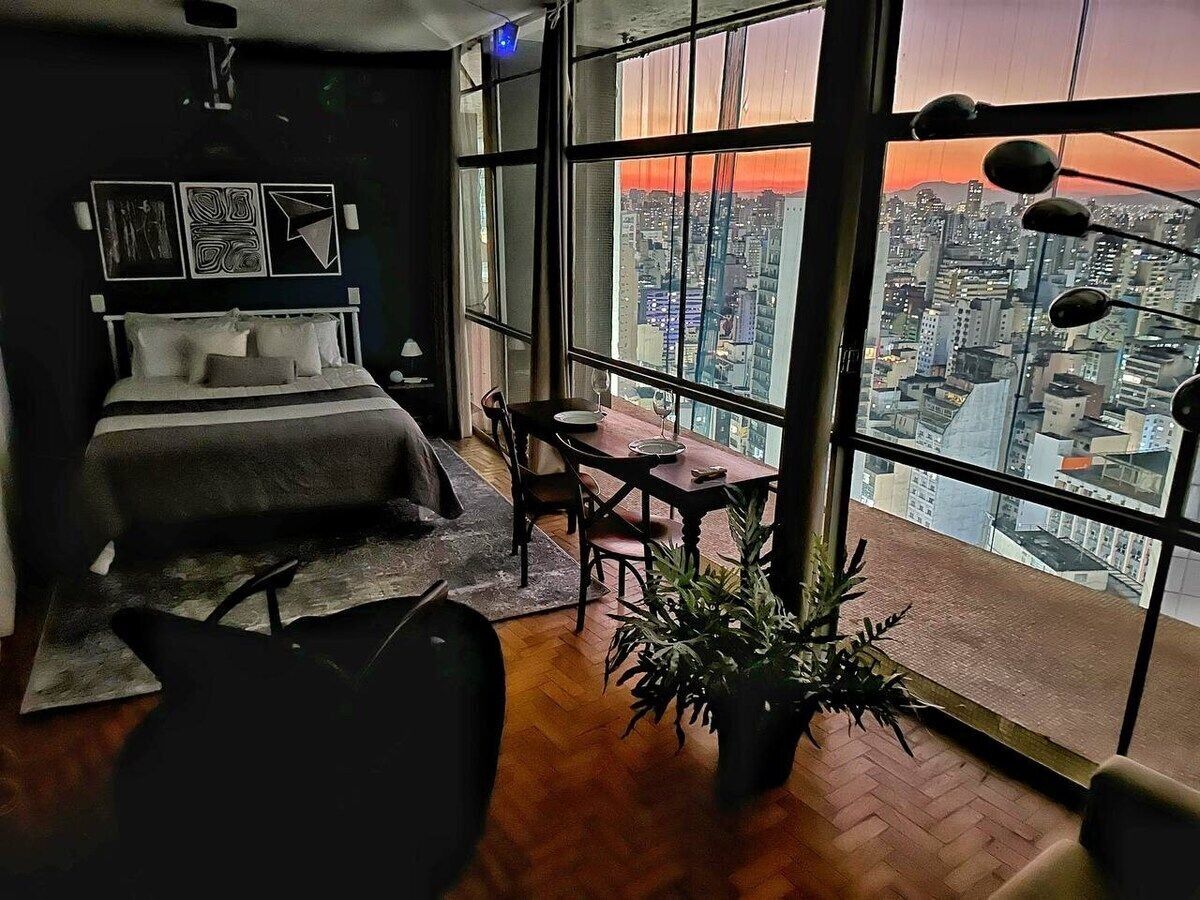 A bedroom with a large window, a bed, a dining table, and a potted plant.