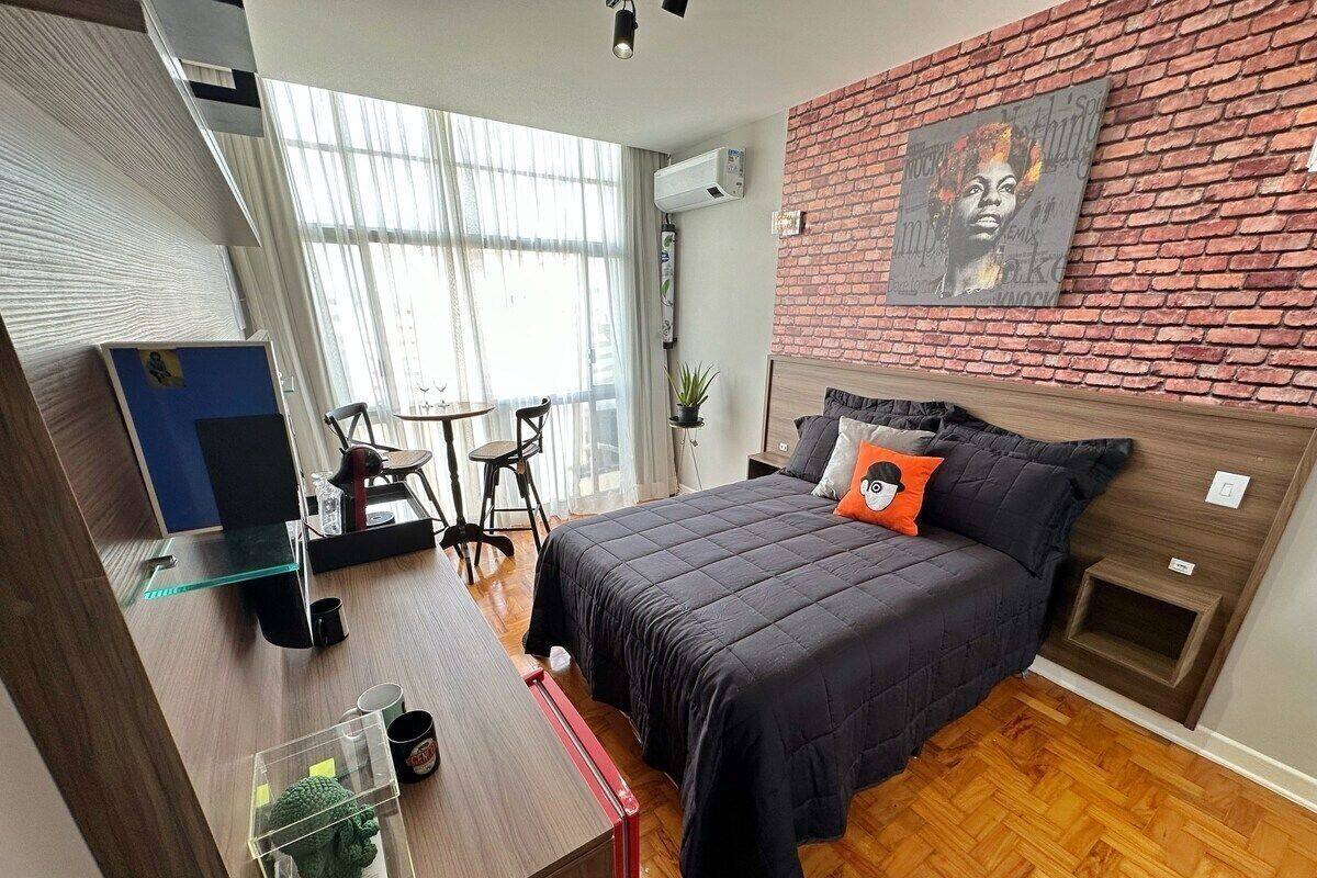 Une chambre moderne avec un mur en briques, un lit recouvert d’une couette noire et agrémenté d’un coussin orange, un petit bureau avec un ordinateur et une fenêtre ornée de rideaux légers.