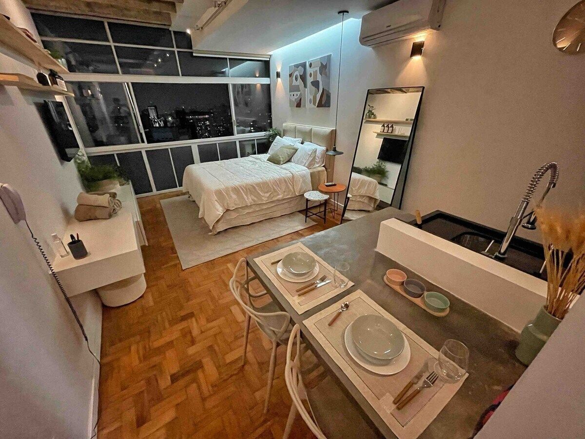Un dormitorio moderno con una cama grande, una mesita de noche y un pequeño escritorio. Se ve una ventana con vistas a la ciudad de noche.