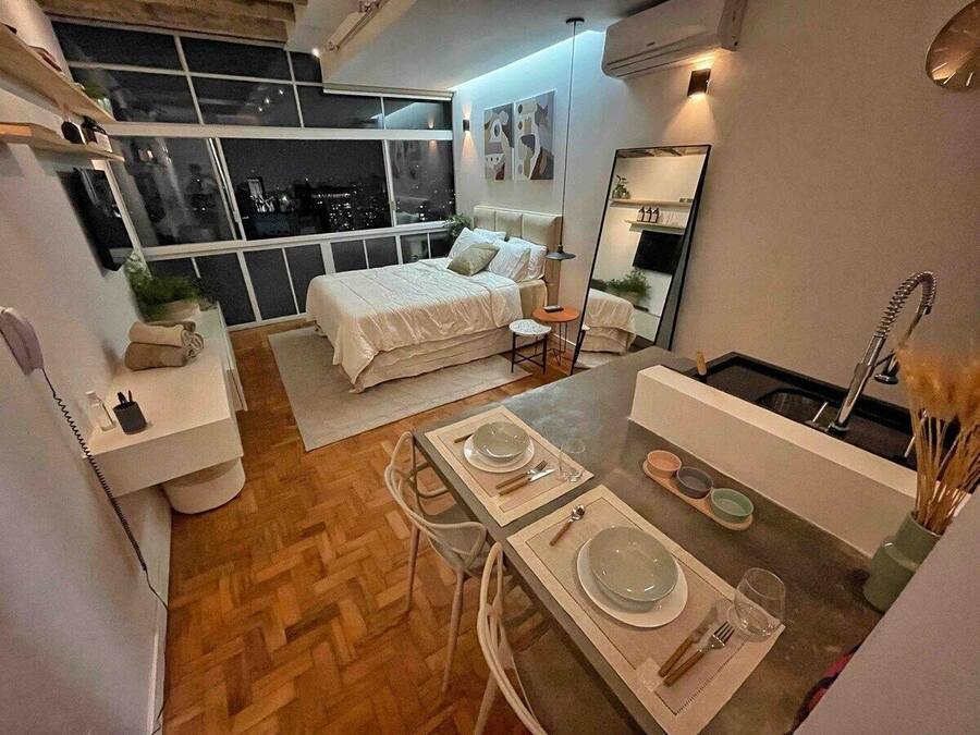 Un dormitorio moderno con una cama grande, una mesita de noche y un pequeño escritorio. Se ve una ventana con vistas a la ciudad de noche.