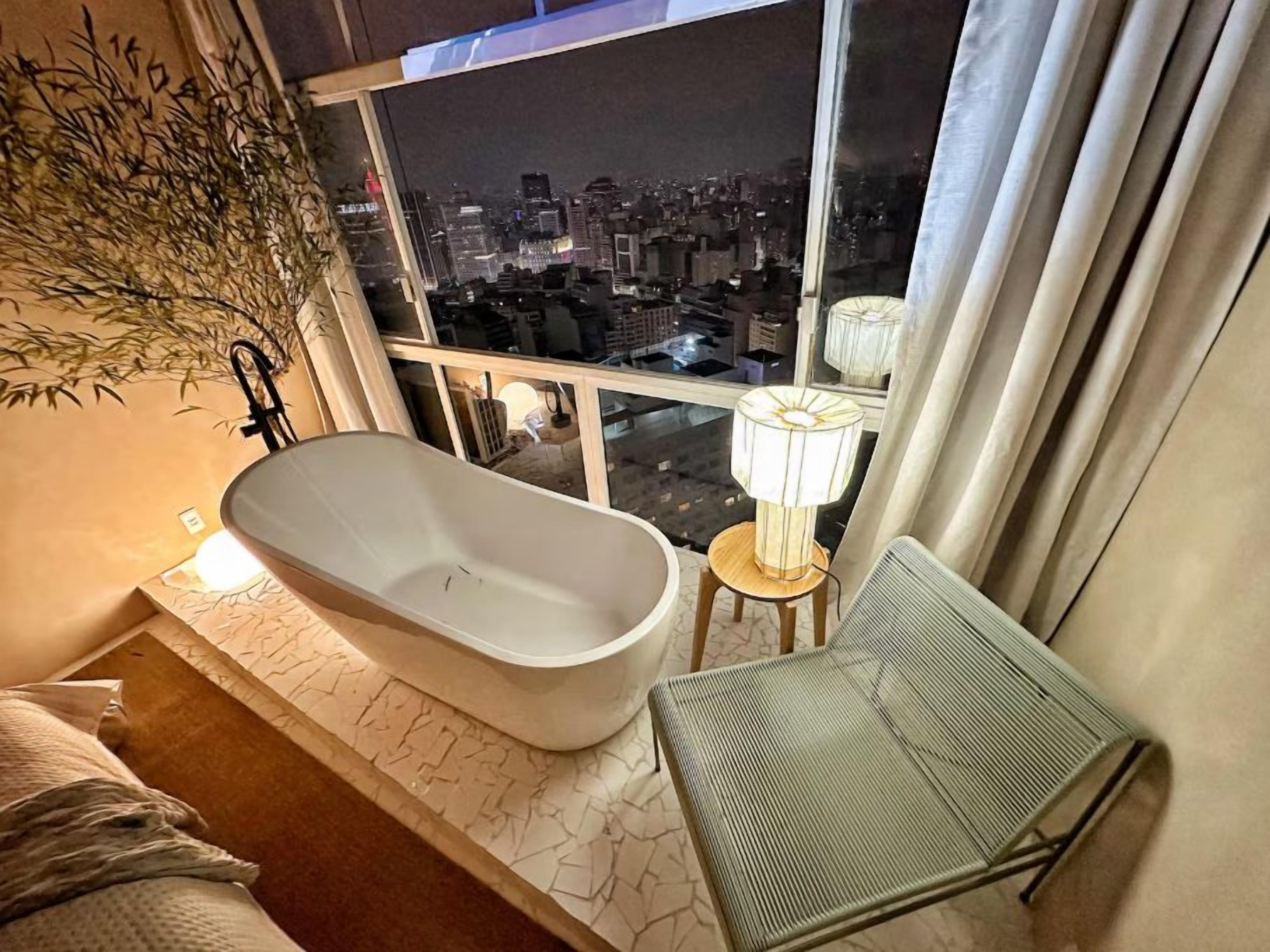 A modern bathroom with a freestanding bathtub, a small stool, and a chair. A large window offers a view of a cityscape at night.