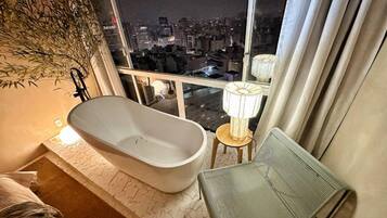 A modern bathroom with a freestanding bathtub, a small stool, and a chair. A large window offers a view of a cityscape at night.