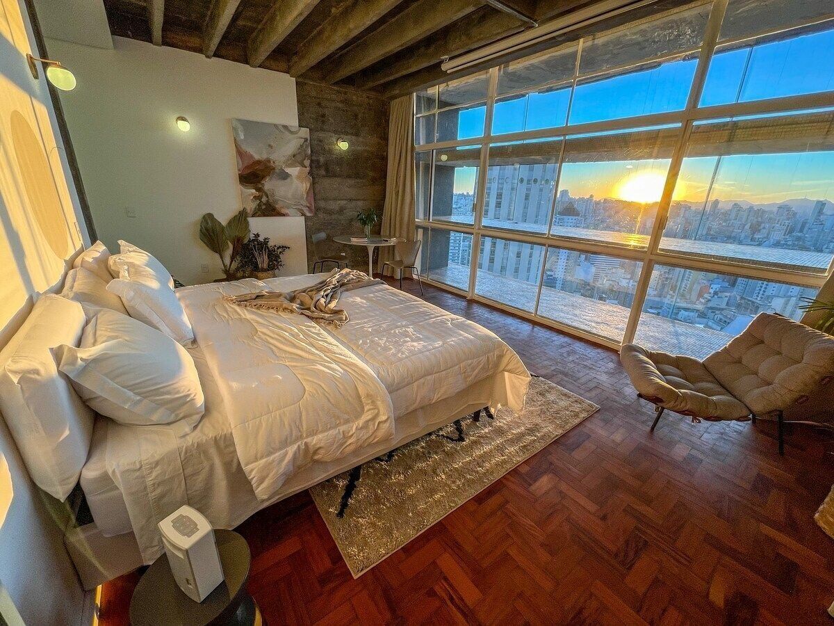 A bedroom with a large bed, a view of the city through a large window, and a modern chair.