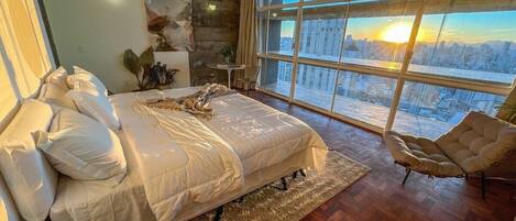 A bedroom with a large bed, a view of the city through a large window, and a modern chair.