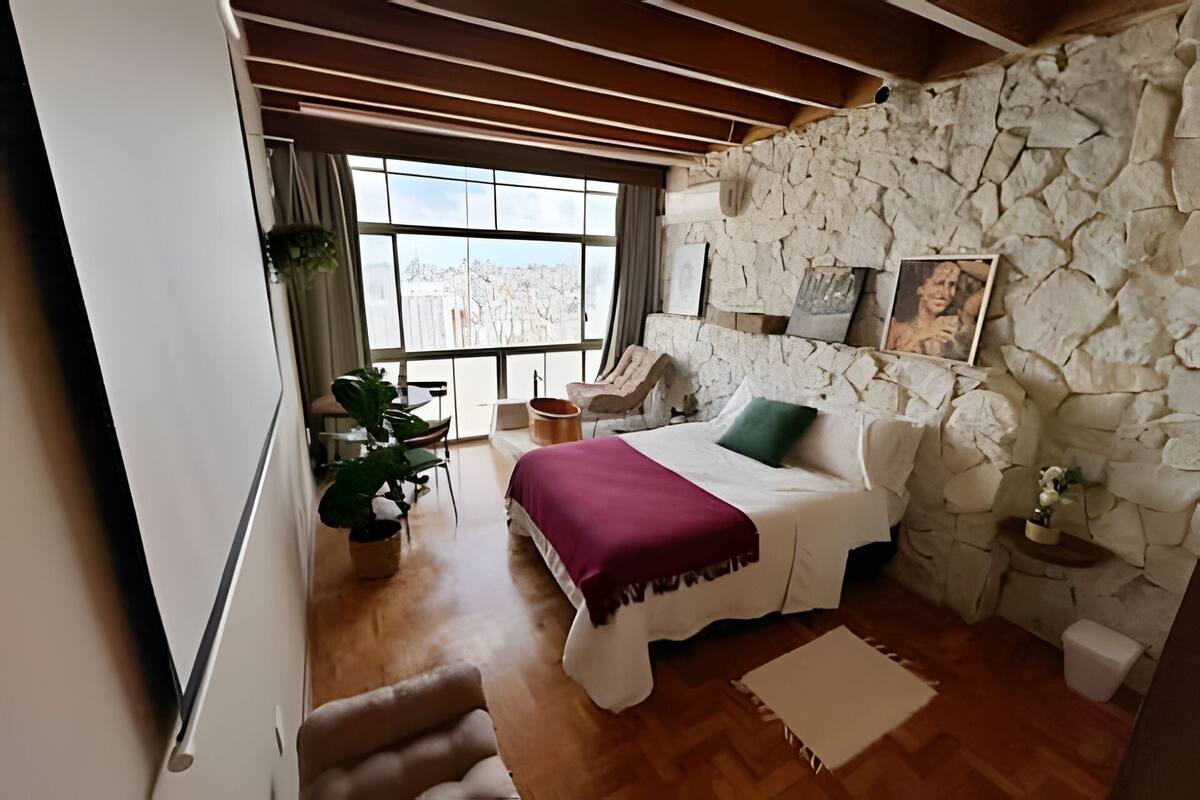 Une chambre avec un mur en pierre, un lit avec un oreiller vert, une fenêtre donnant sur un beau paysage et une plante.