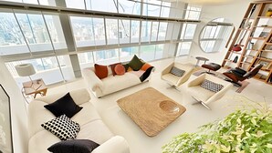 A spacious living room with a large window, a sofa set, a coffee table, a chair, a bookshelf, and a potted plant. - Vem pro Copan (São Paulo)