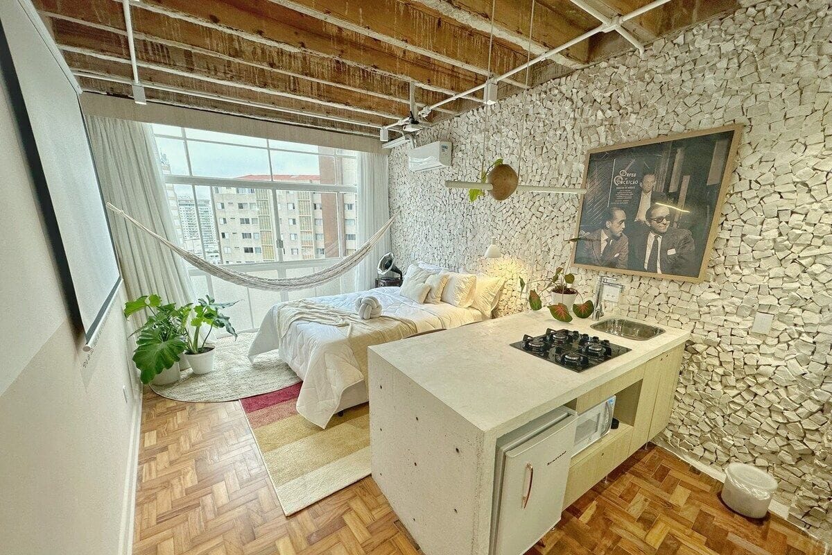 Une chambre moderne avec un grand lit, une petite kitchenette, un mur en pierre et une vue sur les immeubles par la fenêtre.