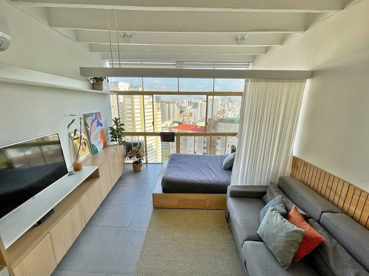 A modern living room with a flat-screen TV, a sofa, a bed, and a balcony with a city view.