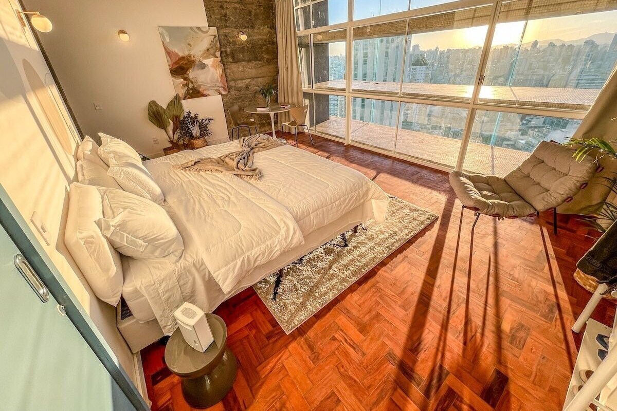 Une chambre dotée d’un grand lit, offrant une vue sur le paysage urbain grâce à de grandes fenêtres du sol au plafond, et dont le plancher est en bois disposé en motif chevrons.