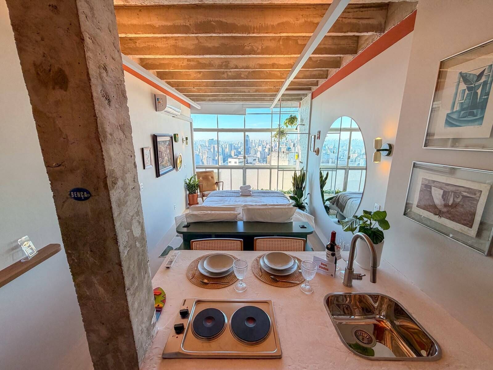 Een moderne keuken met een eethoek, een groot raam met uitzicht op de stad, en een bed met een stadsgezicht zichtbaar door het raam.
