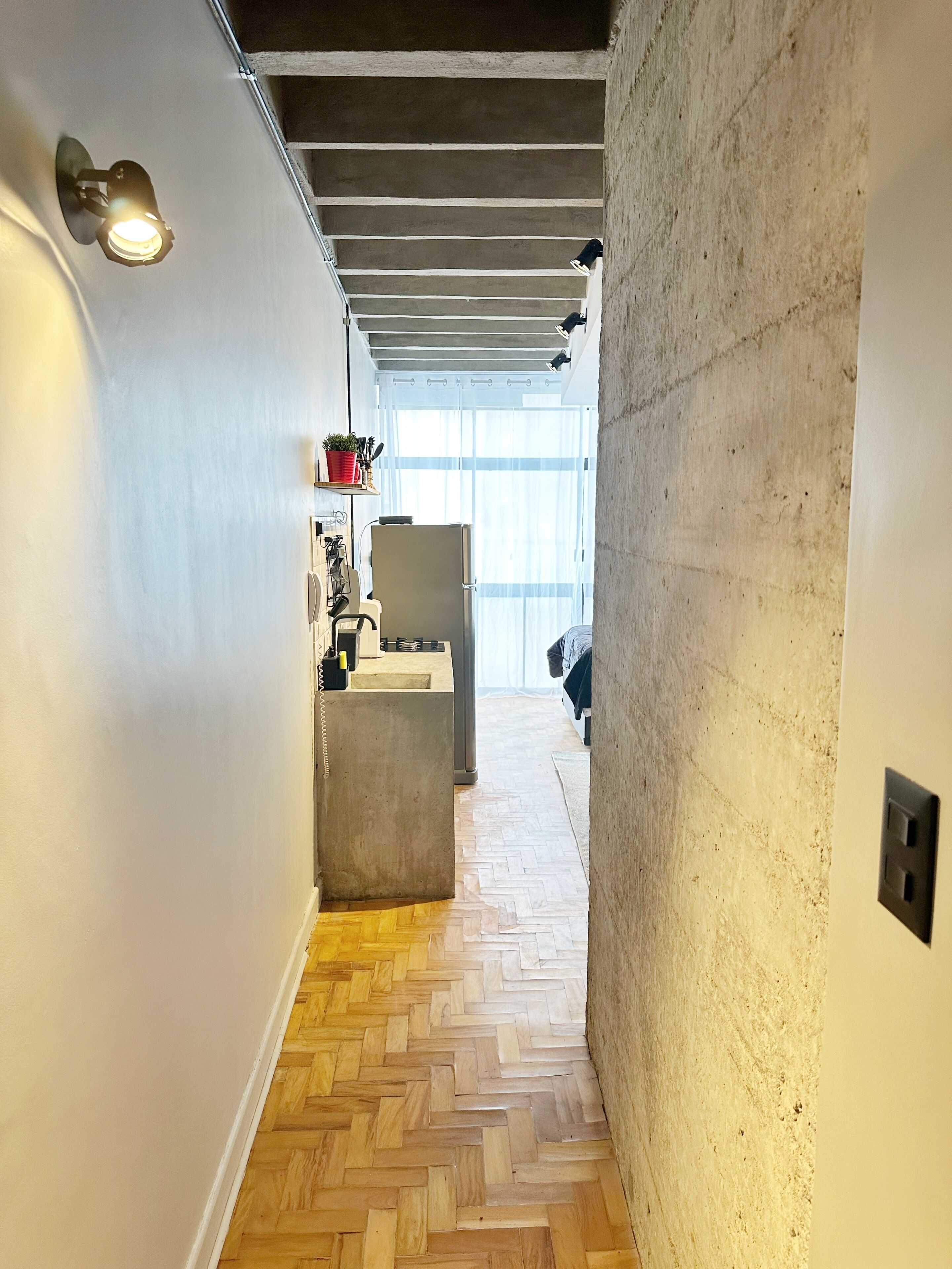 A narrow hallway with a herringbone wooden floor, a concrete wall, and a kitchen area with a refrigerator and a shelf with a plant.