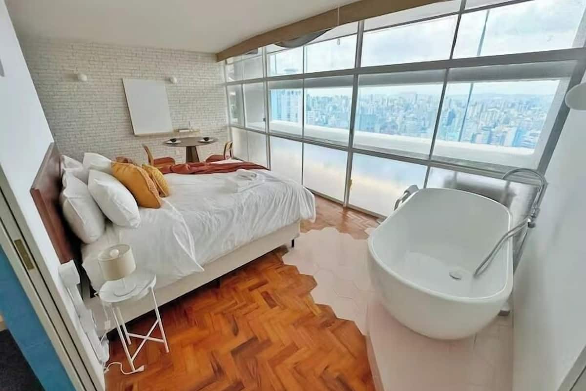 Une chambre moderne avec un grand lit, une baignoire blanche et une vue sur la ville grâce à de grandes fenêtres.
