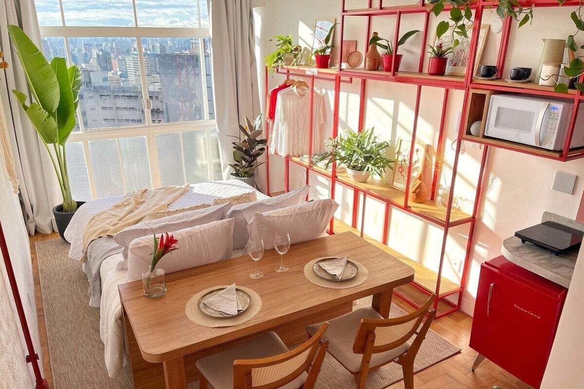 Une chambre confortable avec une grande fenêtre, un coin repas avec une table dressée pour deux personnes, et une étagère rouge où sont disposées des plantes et des objets personnels.