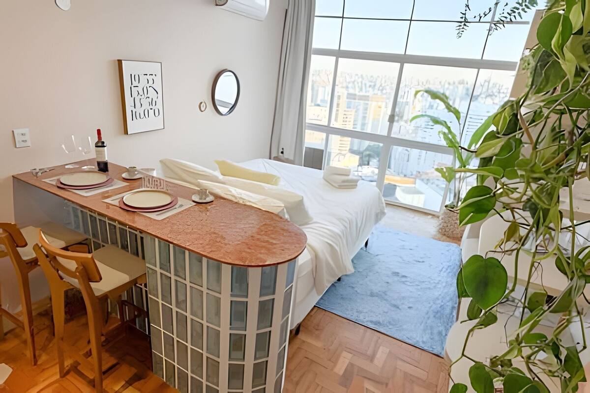 Une chambre d’hôtel moderne dotée d’un coin cuisine, d’un lit avec du linge de maison blanc et d’une grande fenêtre donnant sur la ville.