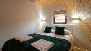A bedroom with a sloped ceiling, a window with blinds, a bed with green and white bedding, and two wall-mounted lights. - 2 Cosy Cabin at 15th C Pub (Wendens Ambo)