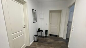 A hallway with wooden flooring, white walls, and two doors. There is a framed picture on the wall and a small table with a plant. - Modern vacation apartment with style & comfort (Ovelgönne)