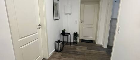 Un pasillo con piso de madera, paredes blancas y dos puertas. Hay un cuadro en la pared y una mesita con una planta.